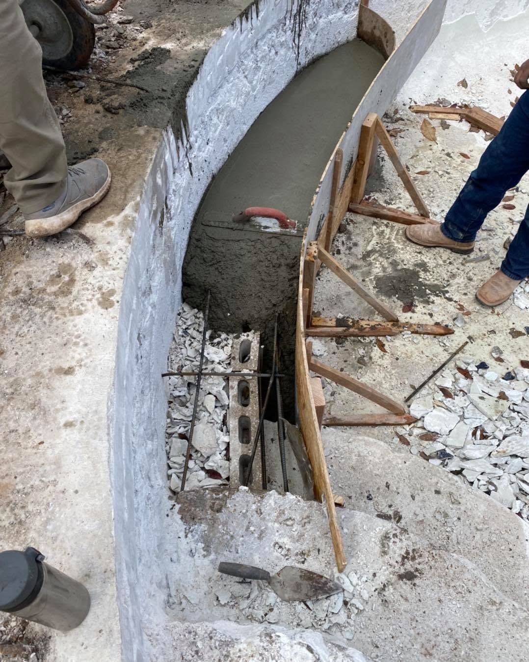 Curved concrete trench with wet cement, rebar, and wooden supports, with workers’ legs visible nearby