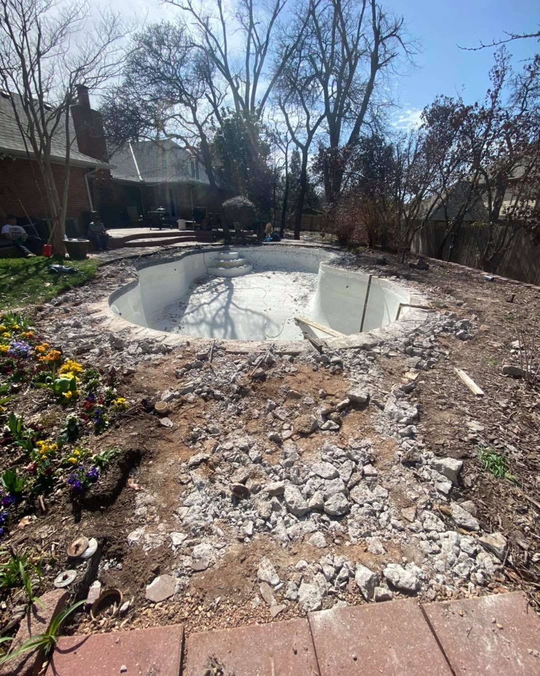 Empty backyard swimming pool under construction, surrounded by dirt, broken concrete, and bare trees.