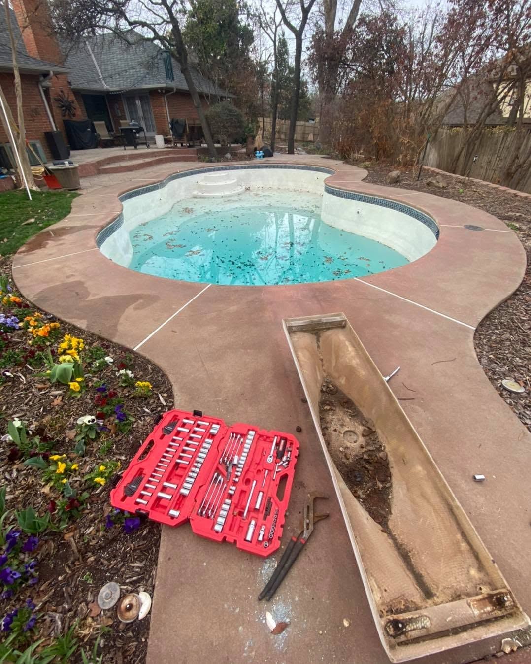 Backyard pool with winter cover removed, tools and red panels on the deck, house and trees in background