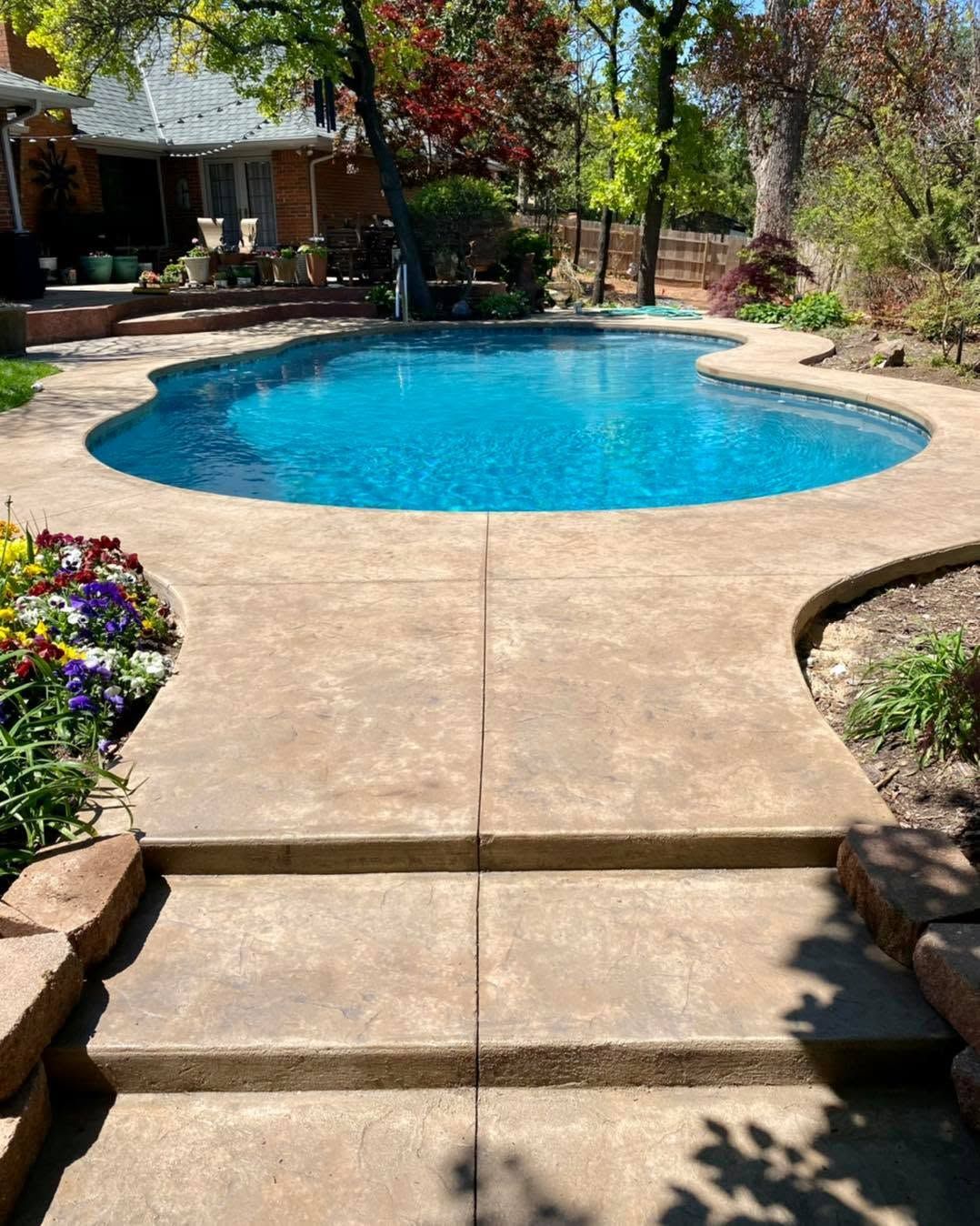 Backyard patio leading to a blue swimming pool, surrounded by trees and flowers.