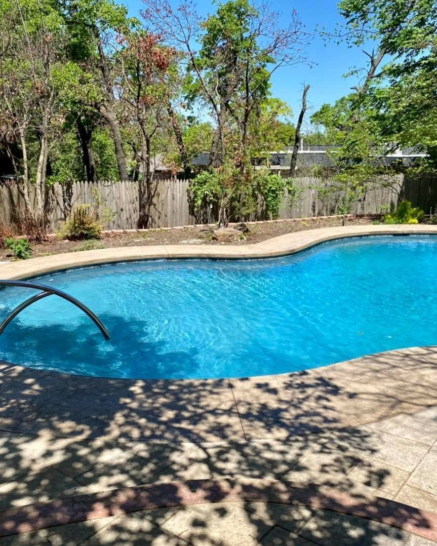 Backyard swimming pool with blue water, stone deck, and trees under a sunny sky