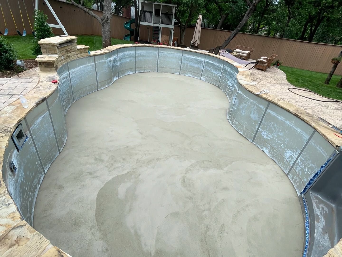 Empty in-ground pool with gray liner and cloudy water, viewed from above in a backyard.