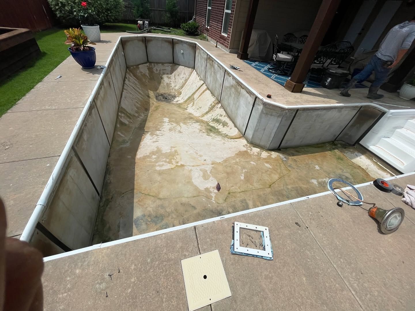 Empty backyard swimming pool under repair, lined with white panels and surrounded by concrete patio.