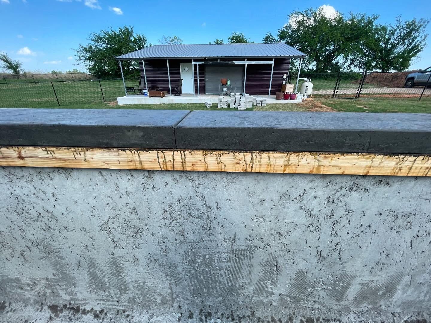 Small house behind a concrete wall, with a road in front and a grassy yard under a blue sky.