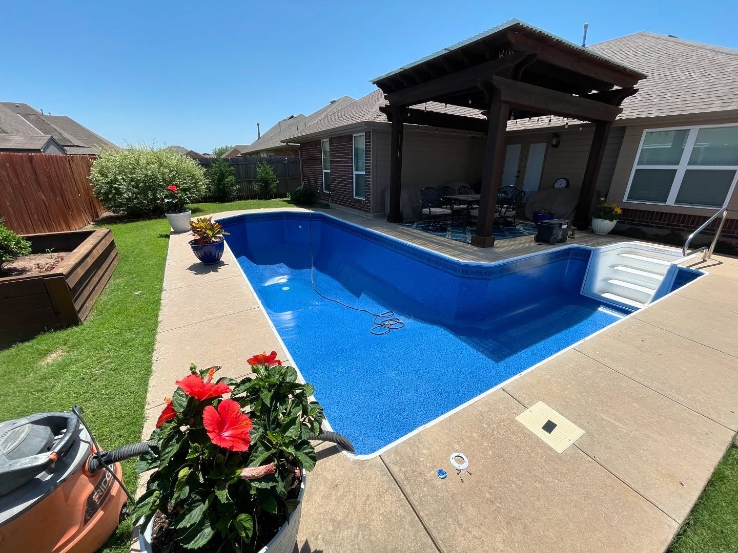 Backyard patio with a blue swimming pool, covered seating area, and potted flowers in bright sunlight