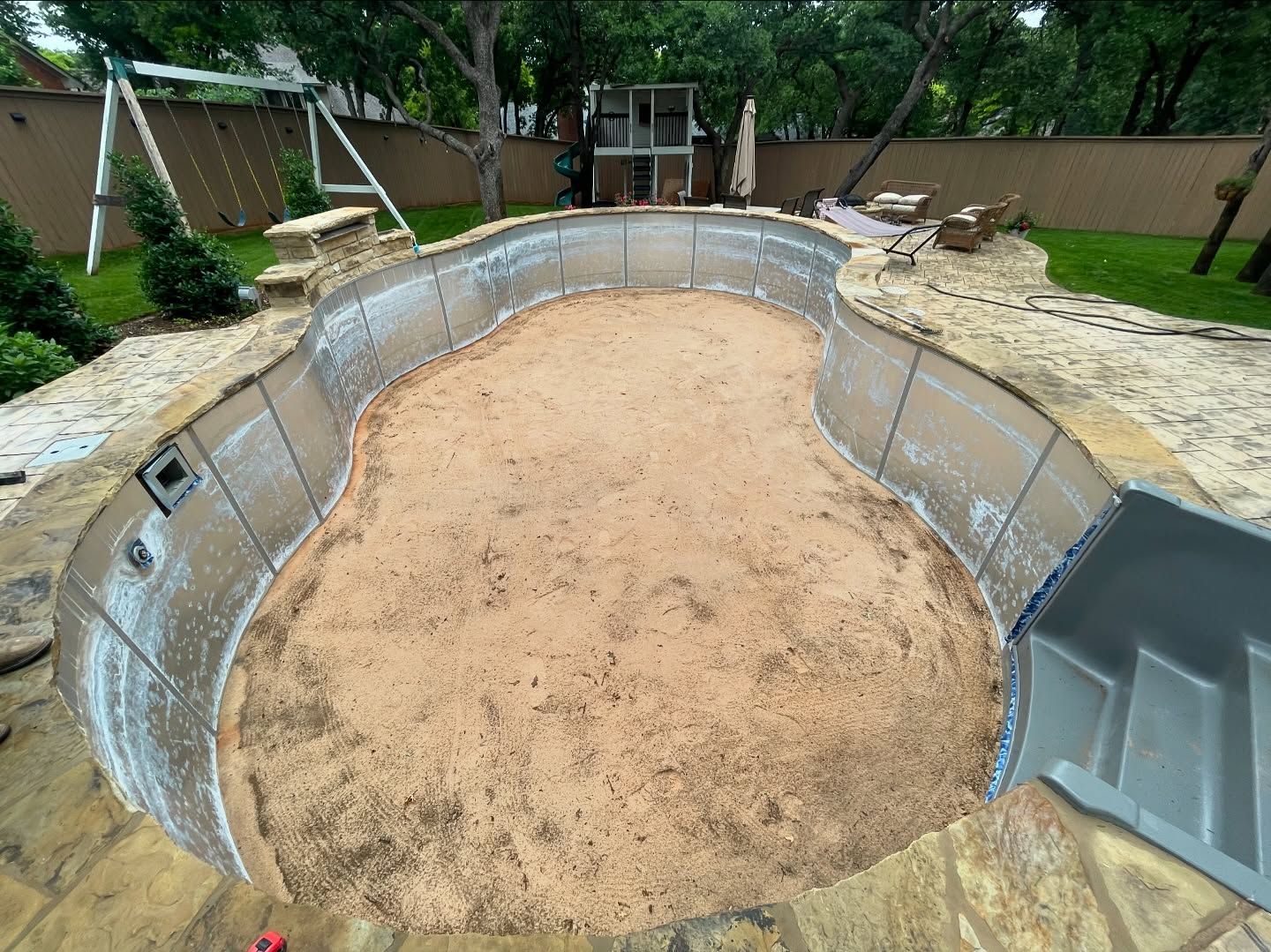 Empty backyard swimming pool under construction, lined with metal walls and sandy floor, with trees and fencing nearby
