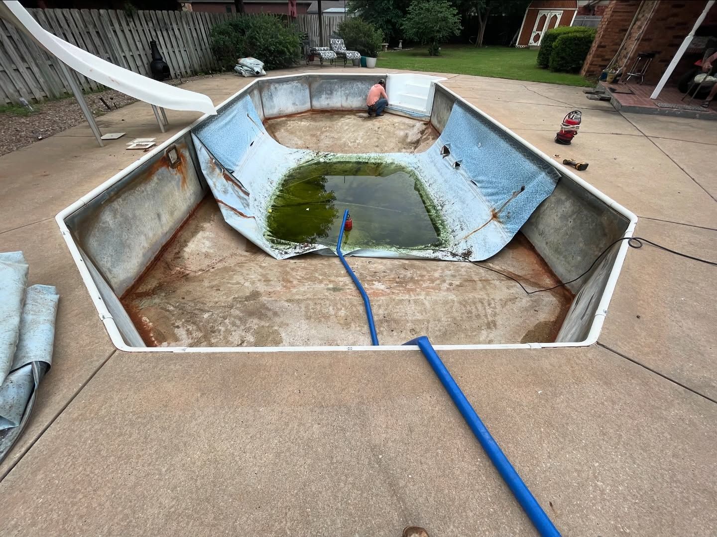 Empty backyard swimming pool with blue hose and debris, drained and in need of cleaning