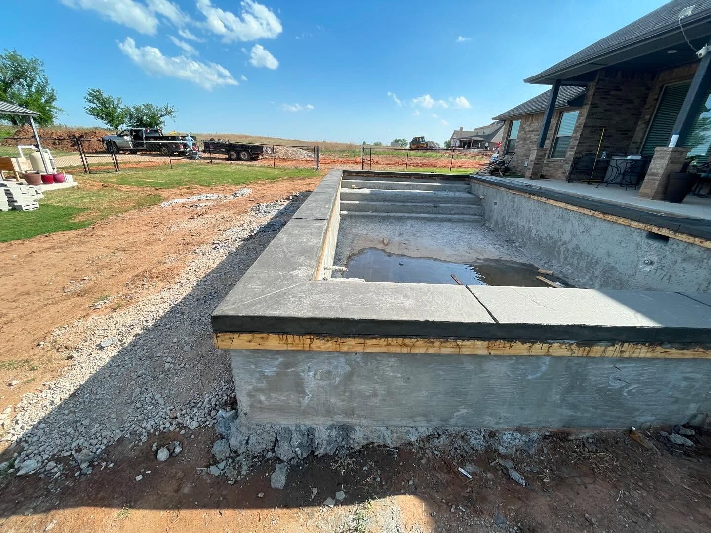 Concrete backyard pool under construction beside a house, with gravel, dirt, and blue sky.