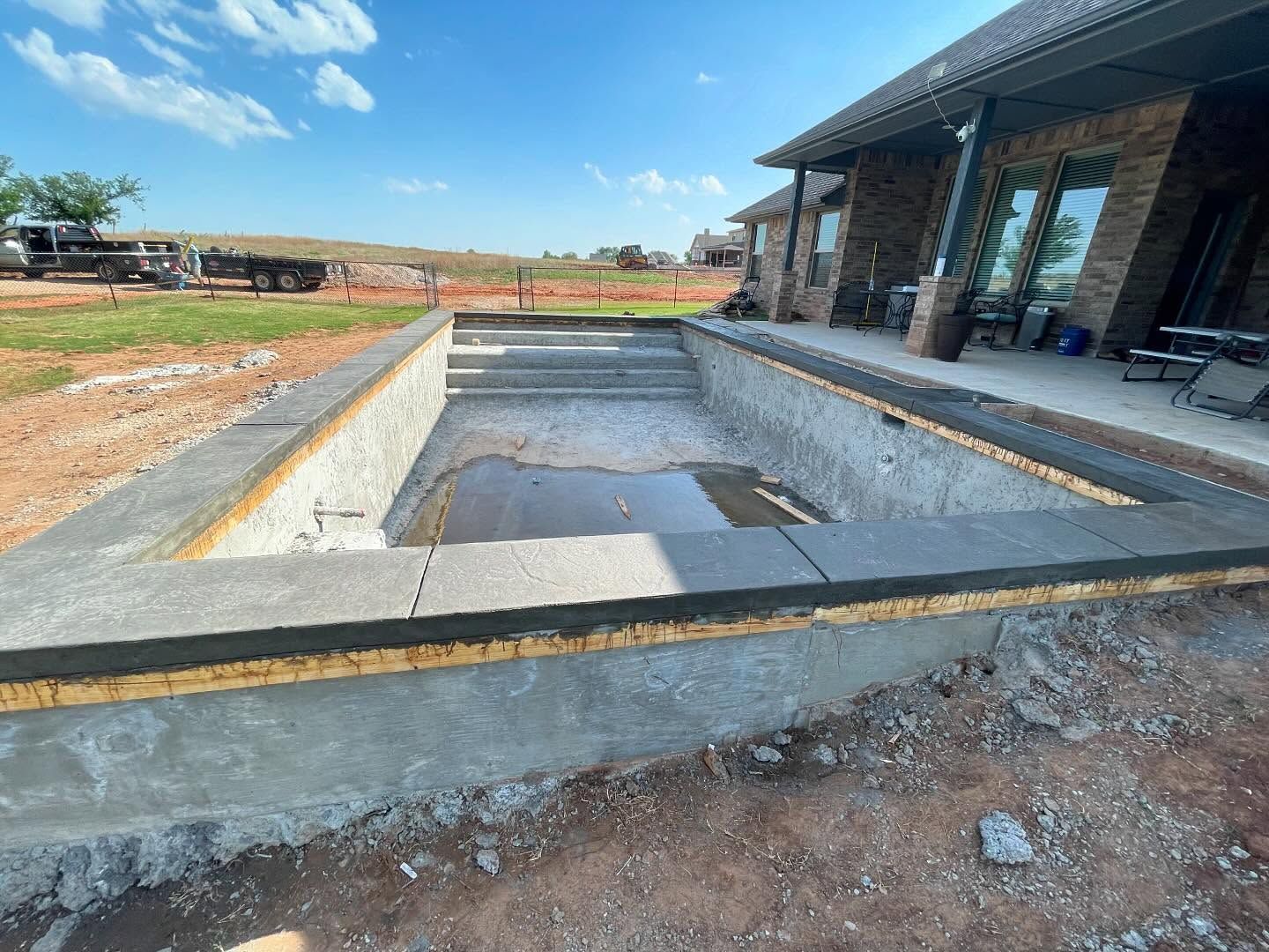 Empty rectangular concrete pool foundation beside a modern house under construction, with open yard and blue sky.