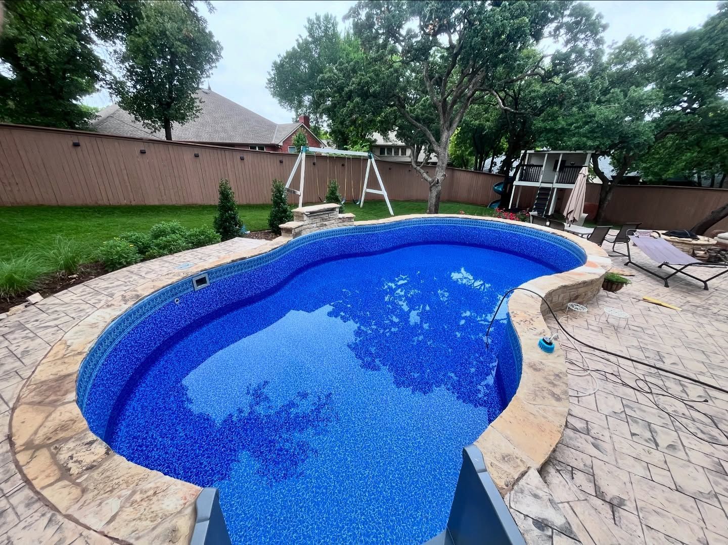 Blue backyard swimming pool with stone patio, lounge chairs, and trees behind a wooden fence