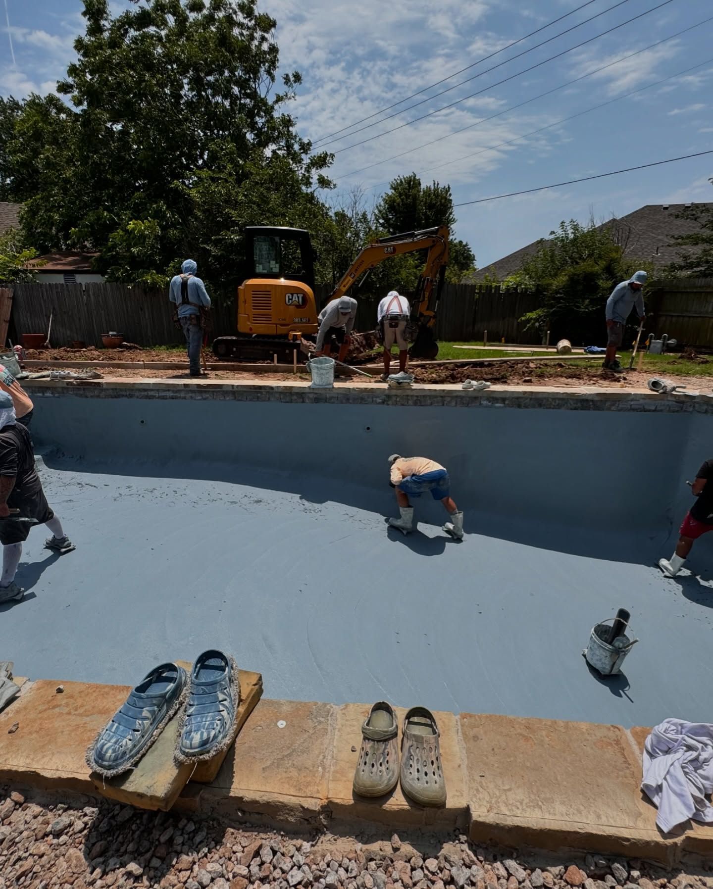 Workers painting a blue pool, with shoes lined up on the edge and a backhoe nearby under a sunny sky
