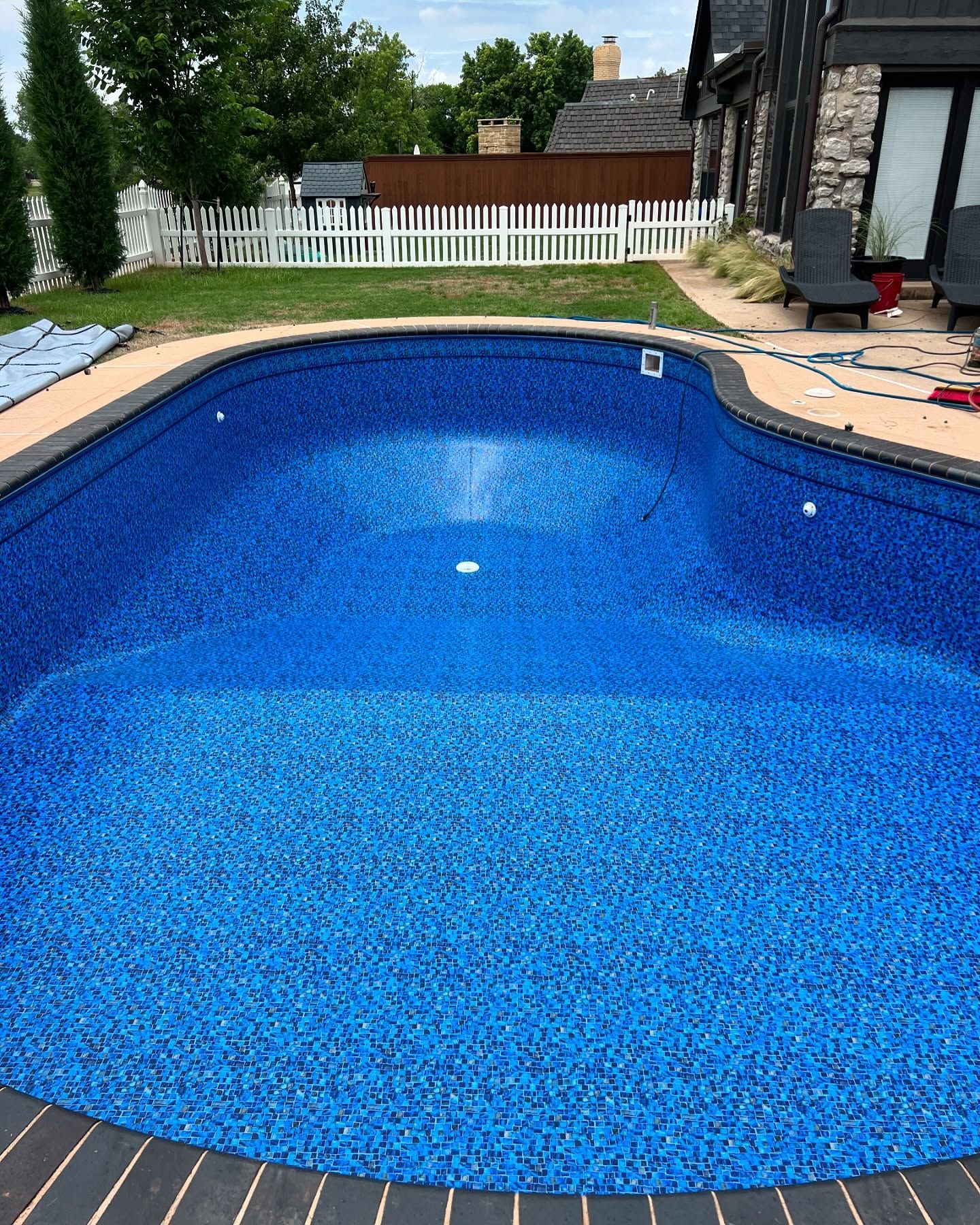 Blue backyard swimming pool beside a house, with white fence and patio furniture nearby