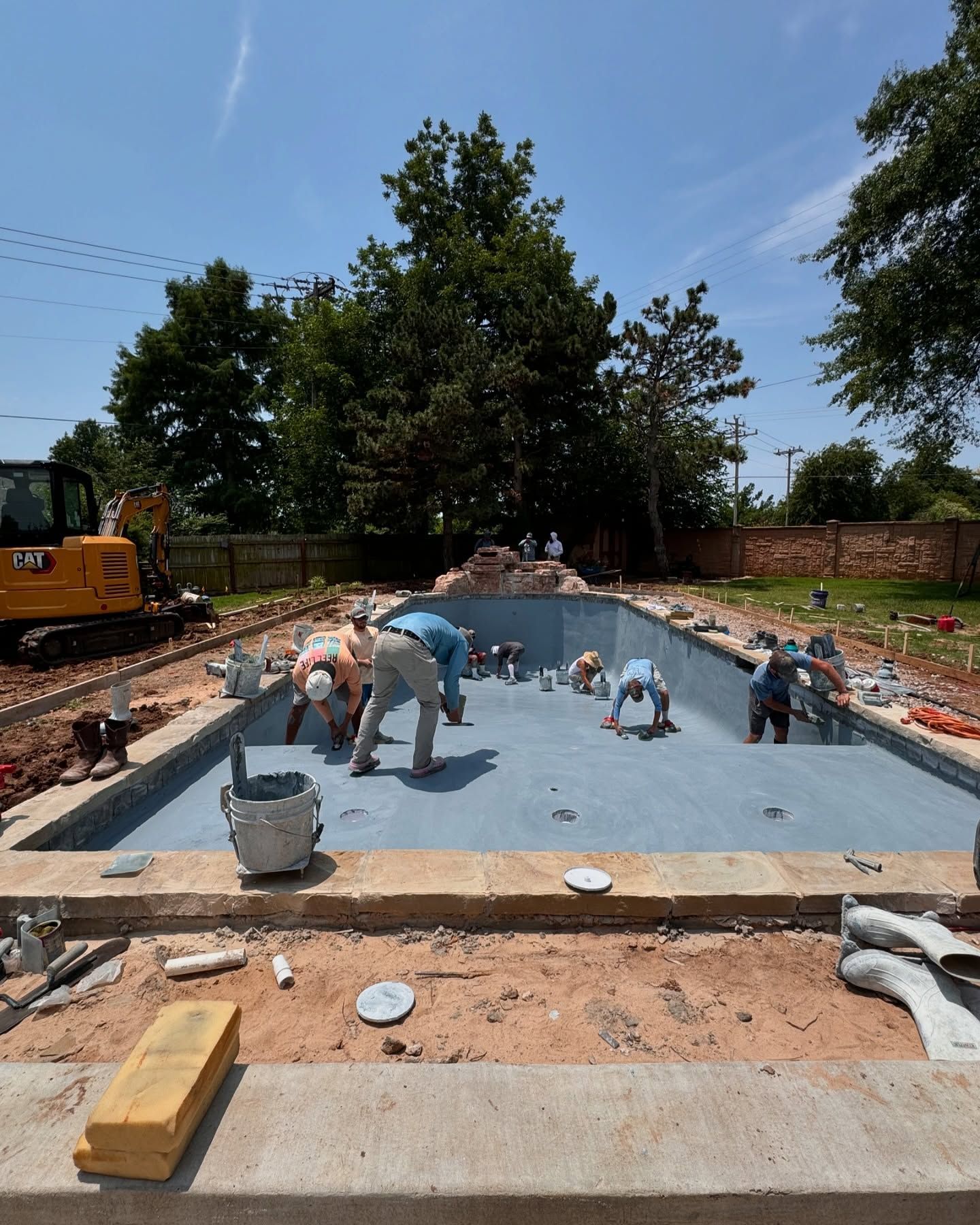 Workers tiling a shallow pool, with blue floor, tools, and construction equipment at a backyard site