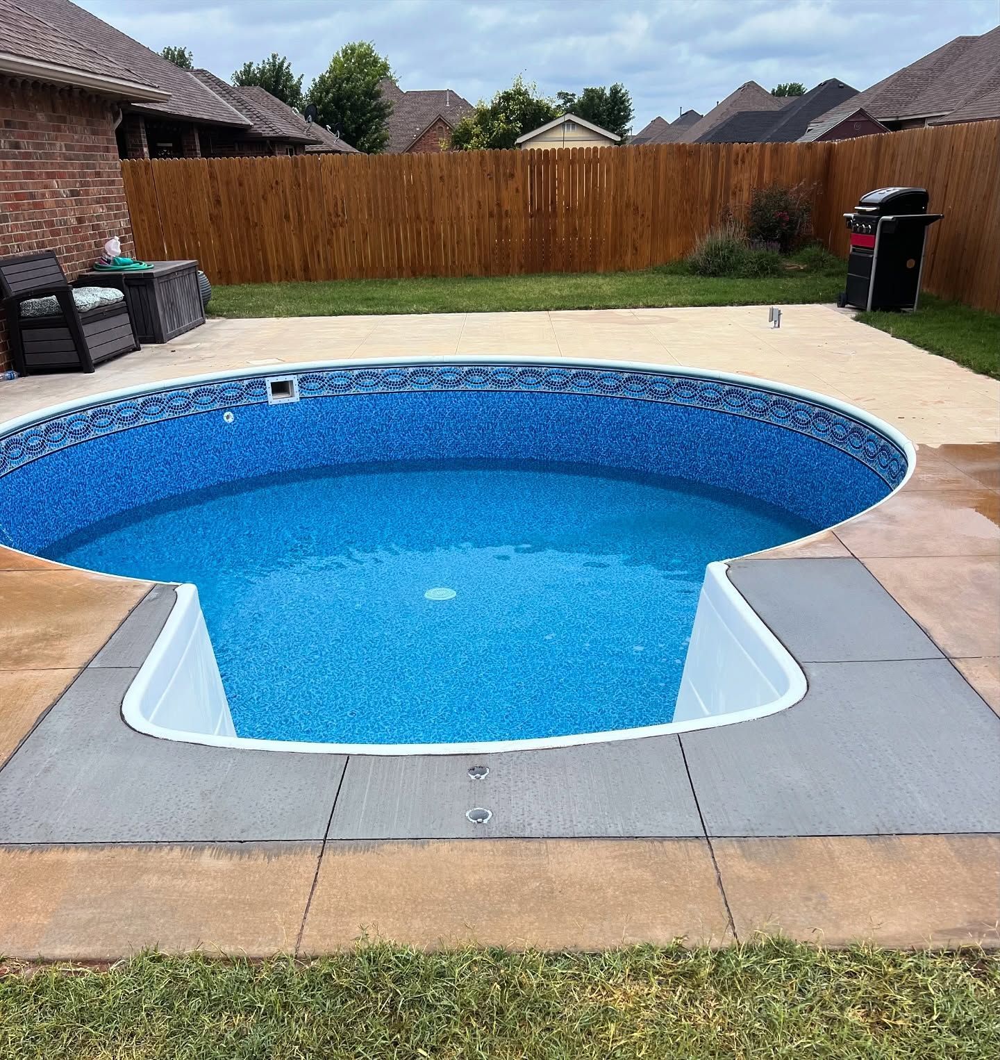 Backyard swimming pool with blue water, gray deck, wooden fence, and patio seating