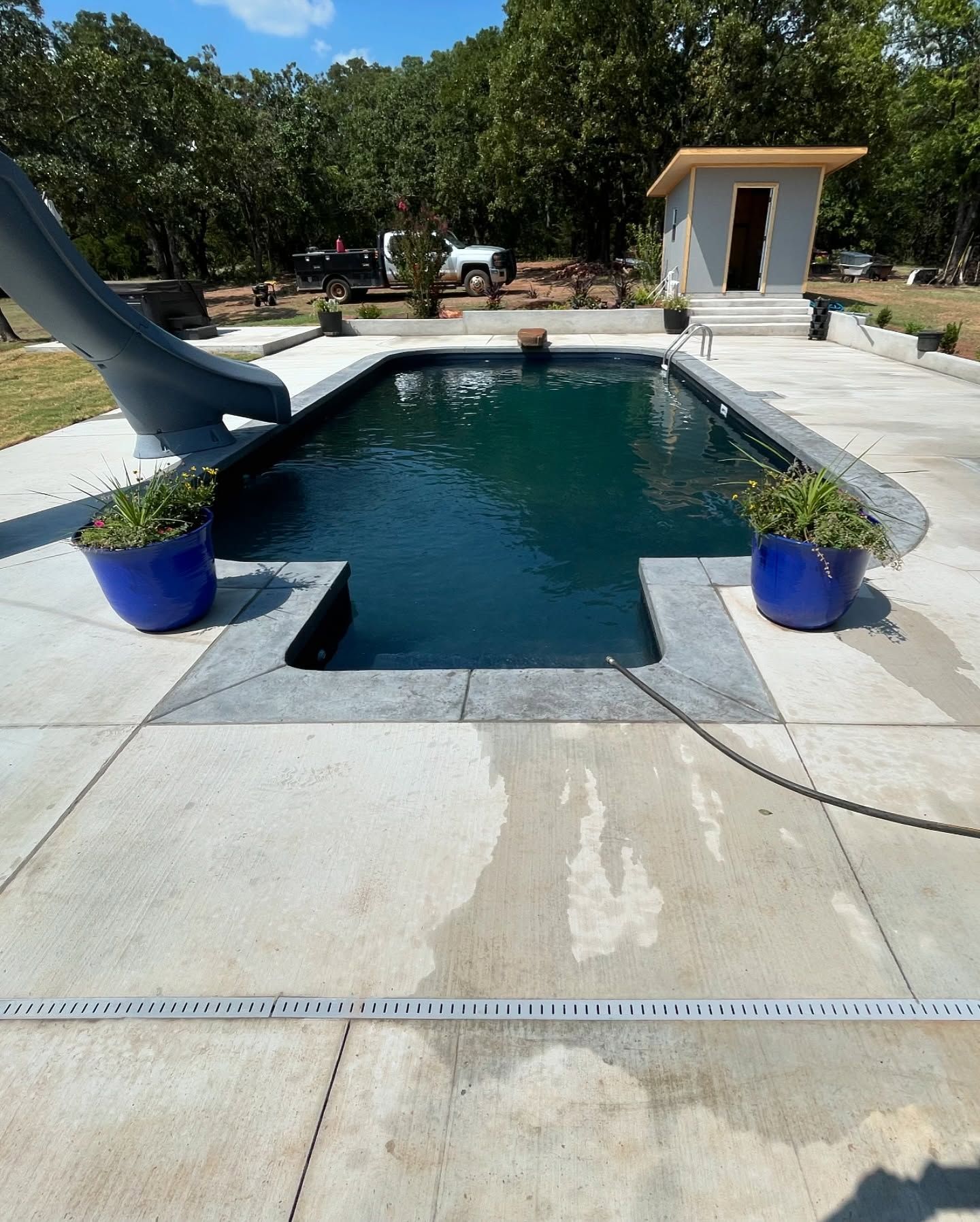 Backyard pool with slide, blue planters, and a small pool house on a sunny day