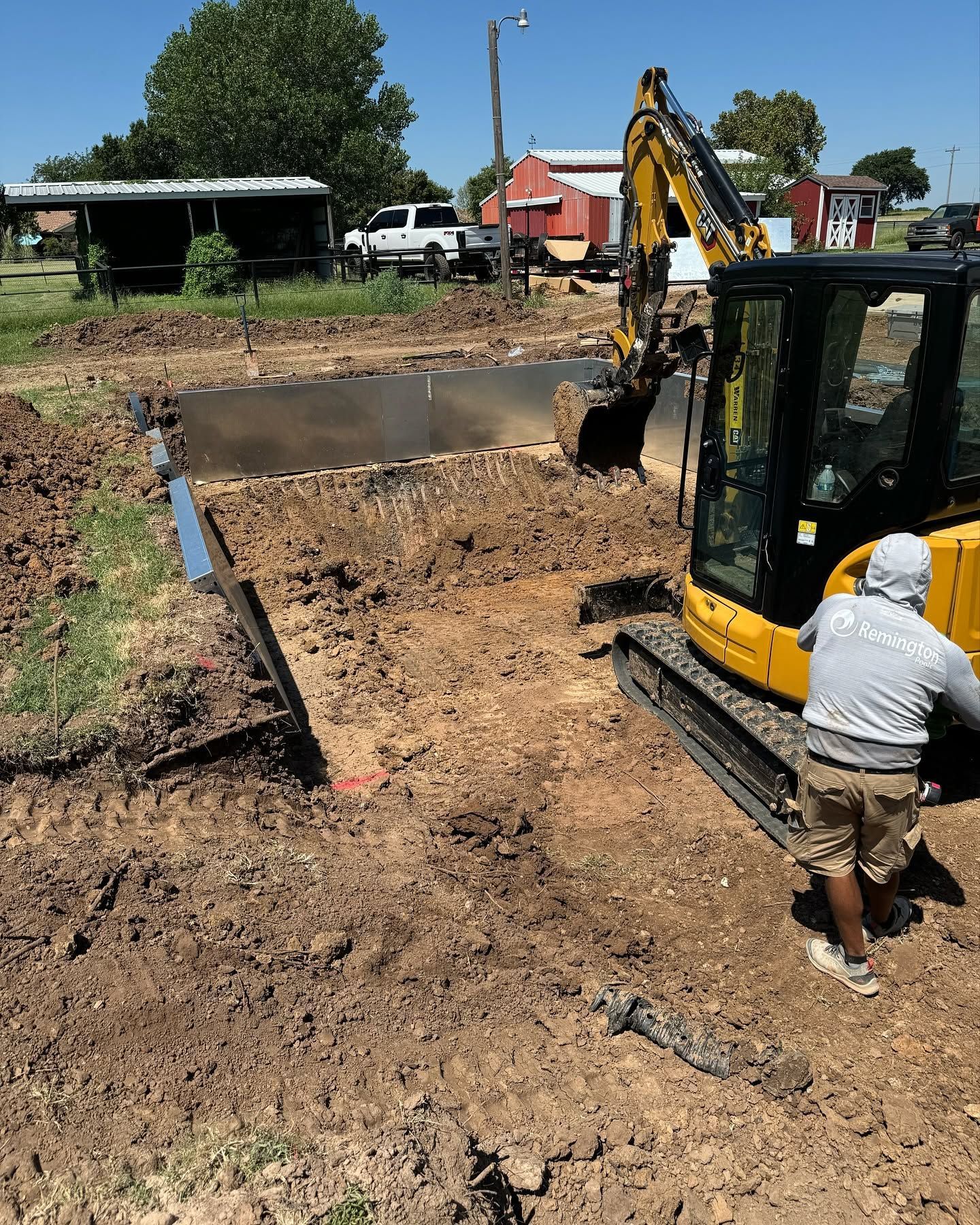 Backhoe excavating dirt beside a concrete foundation, with a worker nearby in a sunny outdoor lot
