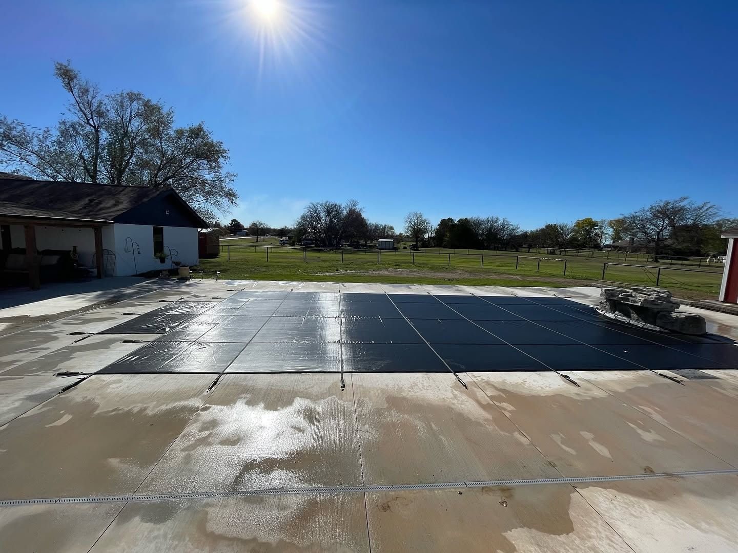 Solar panels laid out on a concrete patio beside a house under a bright blue sky.