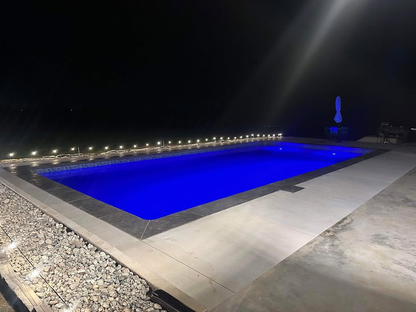 Illuminated blue swimming pool at night with surrounding walkway and distant lights