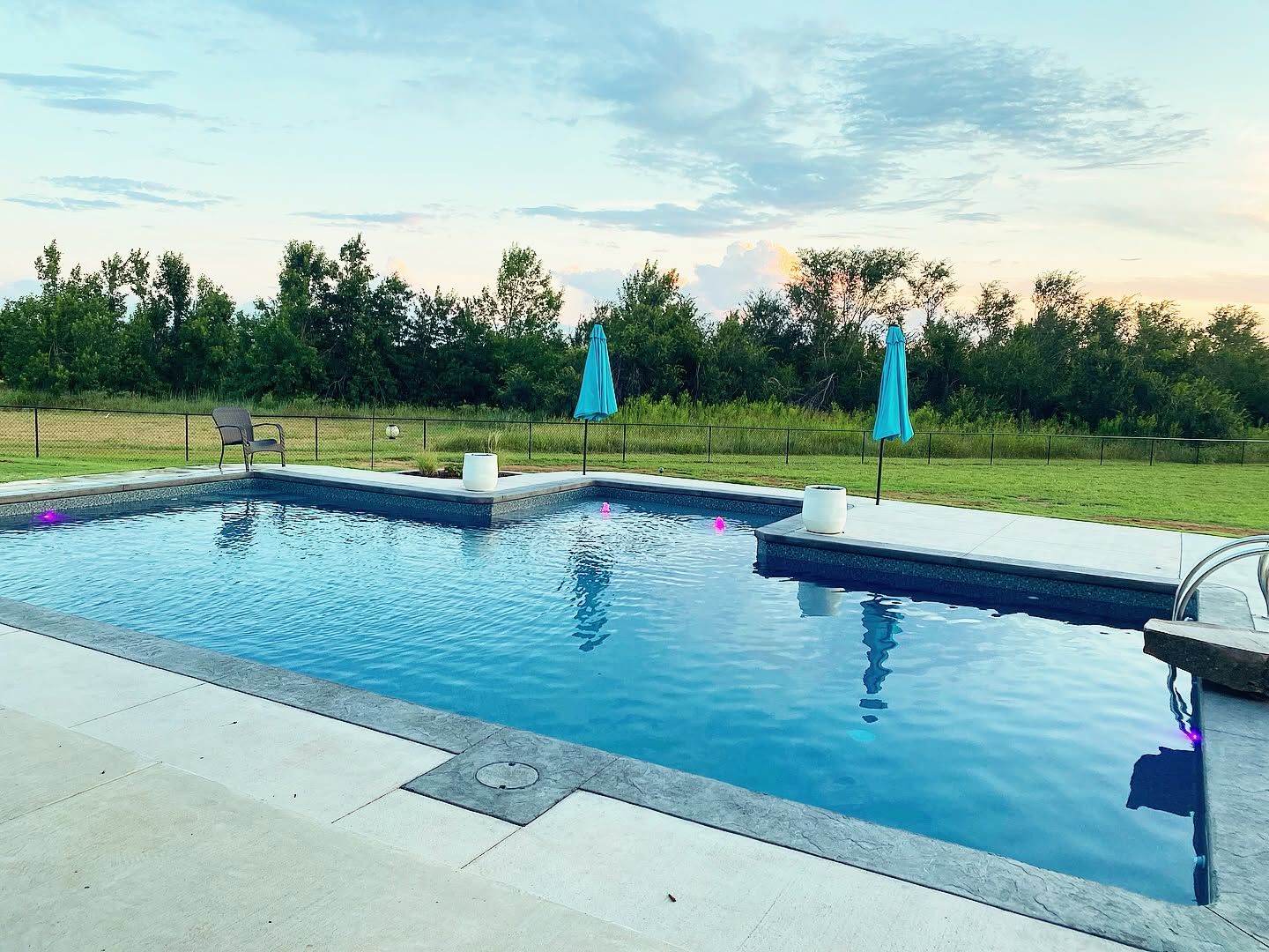 Backyard pool with blue water, turquoise umbrellas, and grassy field at sunset