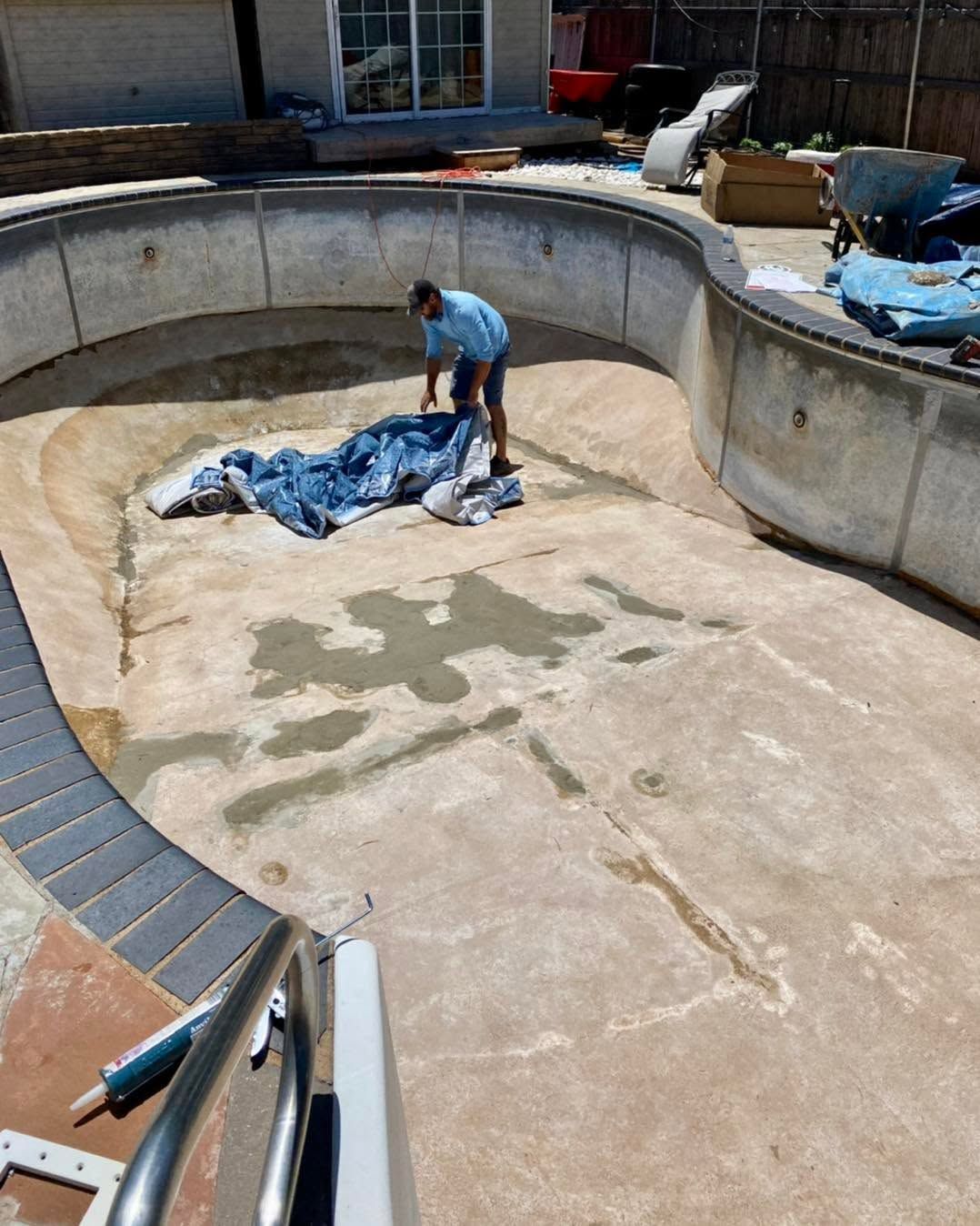 Person cleaning an empty backyard pool under a blue tarp, with a hose and scattered equipment nearby.