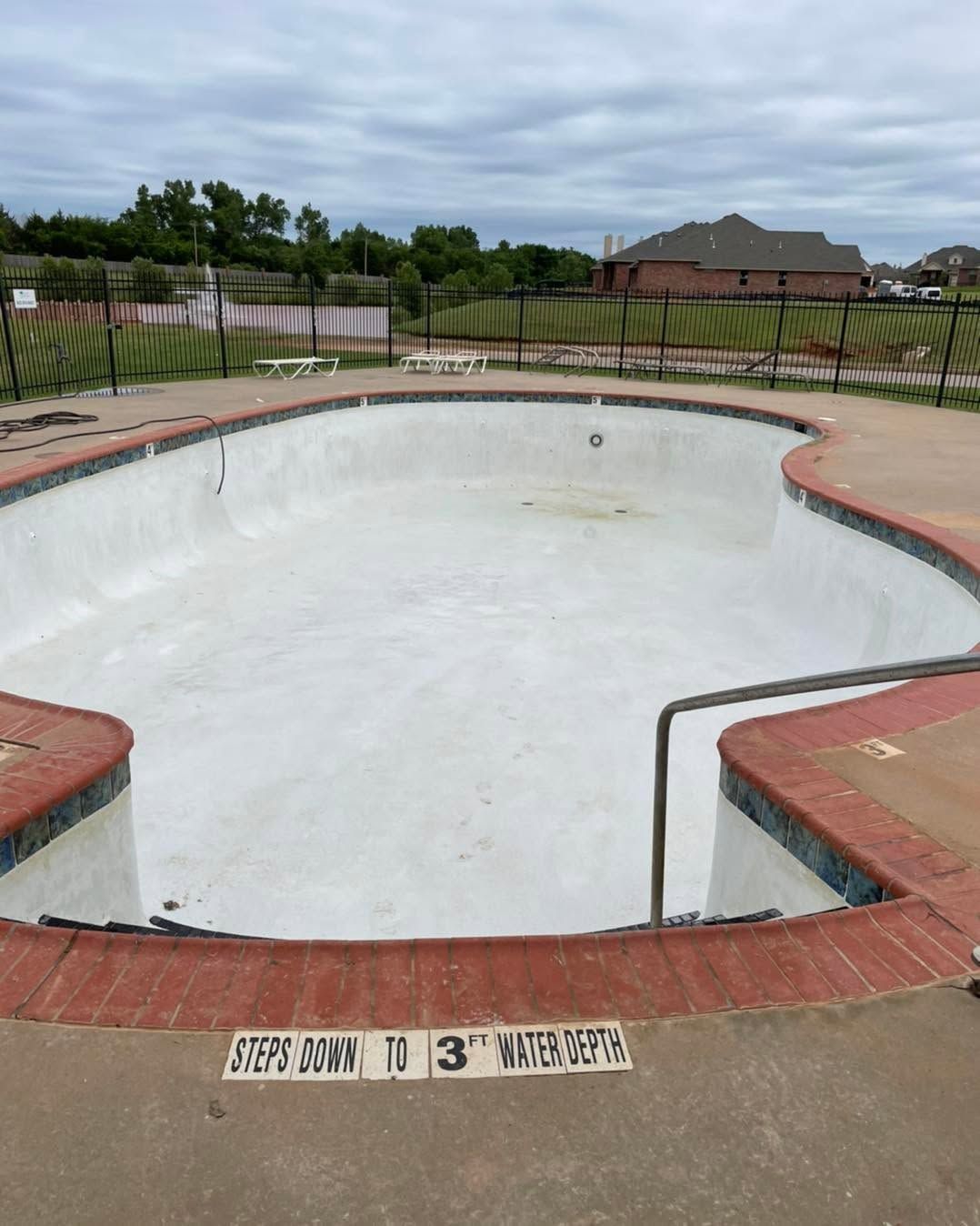 Empty drained swimming pool with white basin, red brick coping, and metal railings under an overcast sky