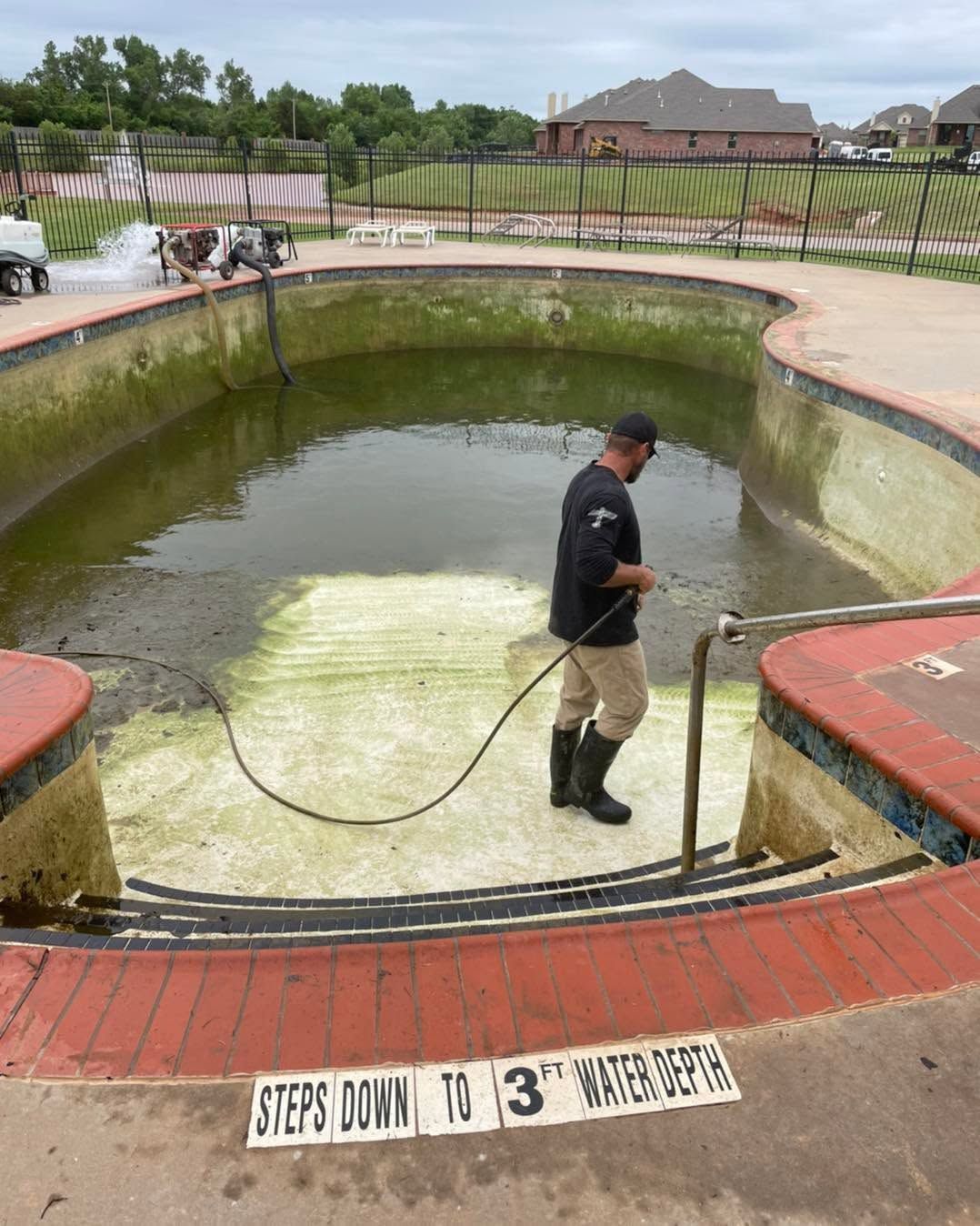 Worker cleaning a drained, algae-lined pool with a hose and broom in a suburban backyard.