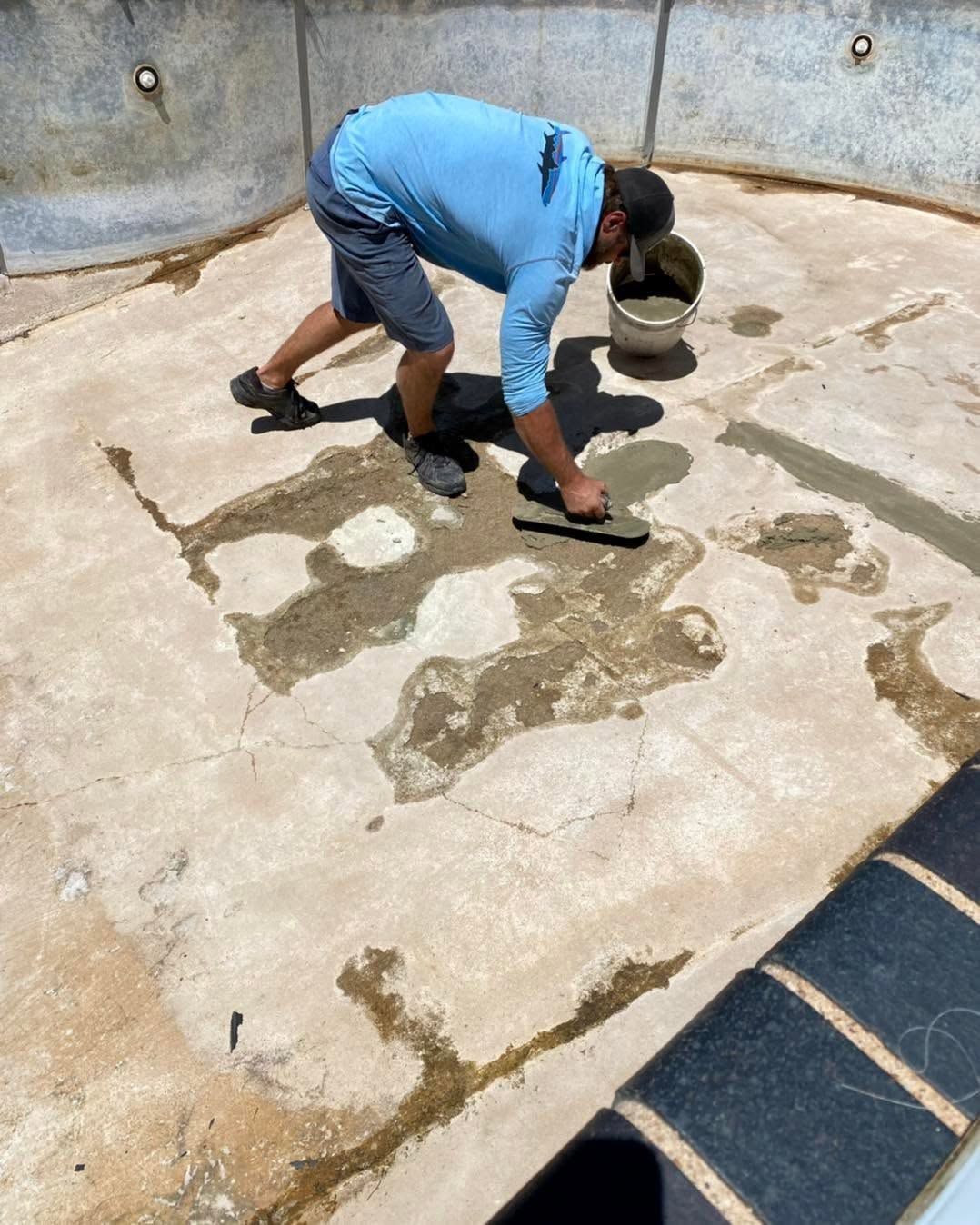Worker kneeling on a concrete surface, applying or patching wet cement near a pool area.