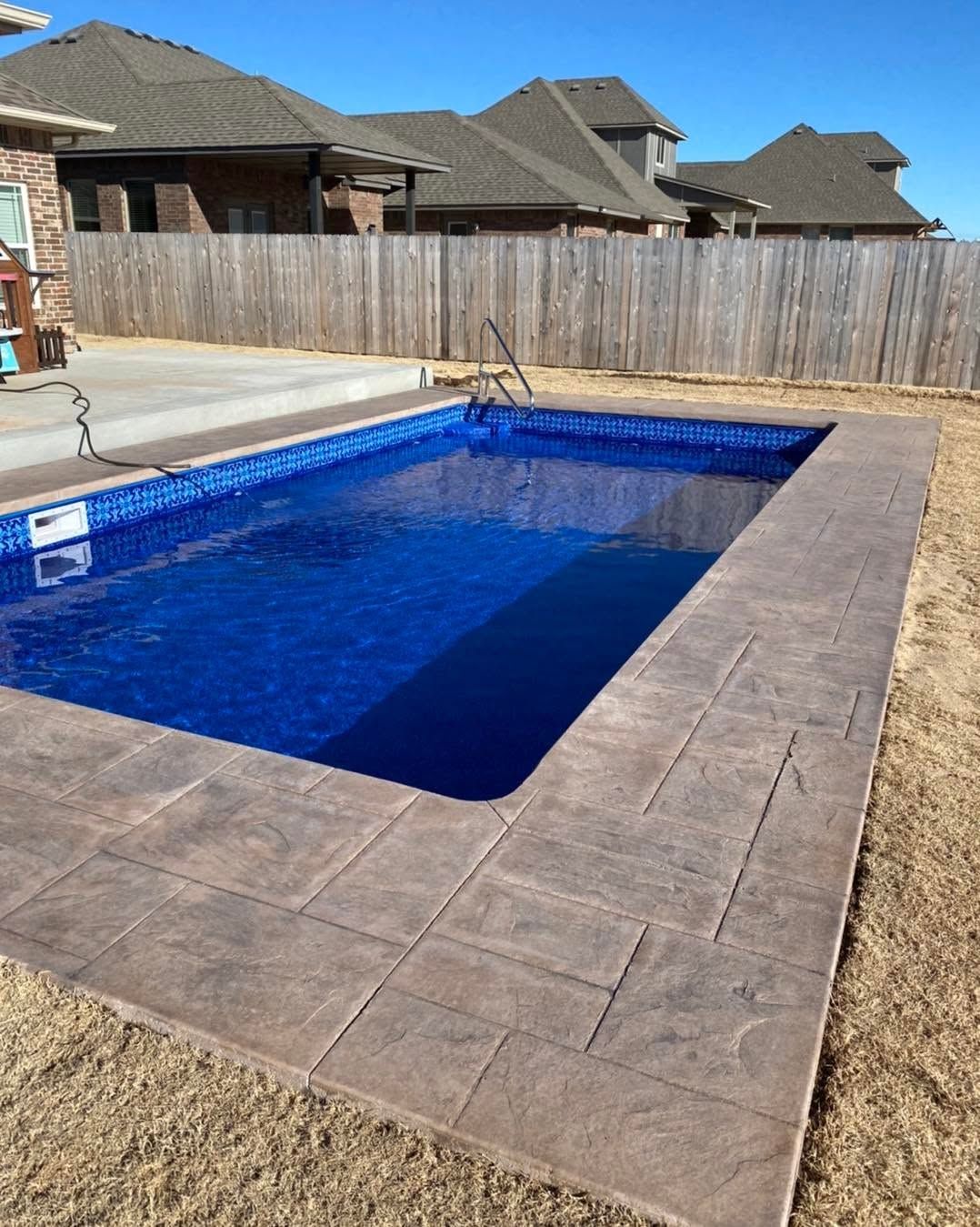 Backyard rectangular pool with blue water and a stone patio, bordered by a wooden fence.