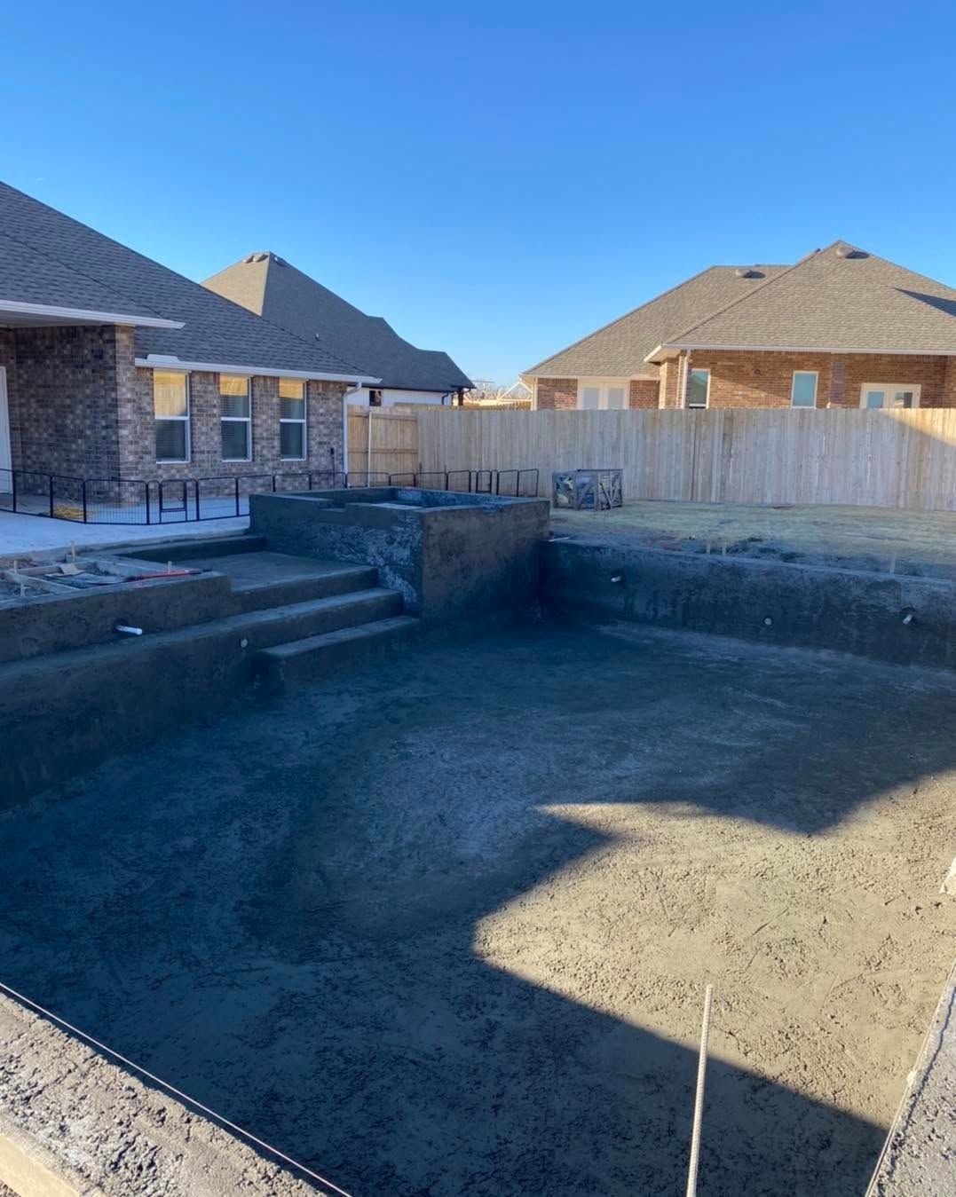 Backyard with an empty rectangular swimming pool under construction beside brick houses and a wooden fence