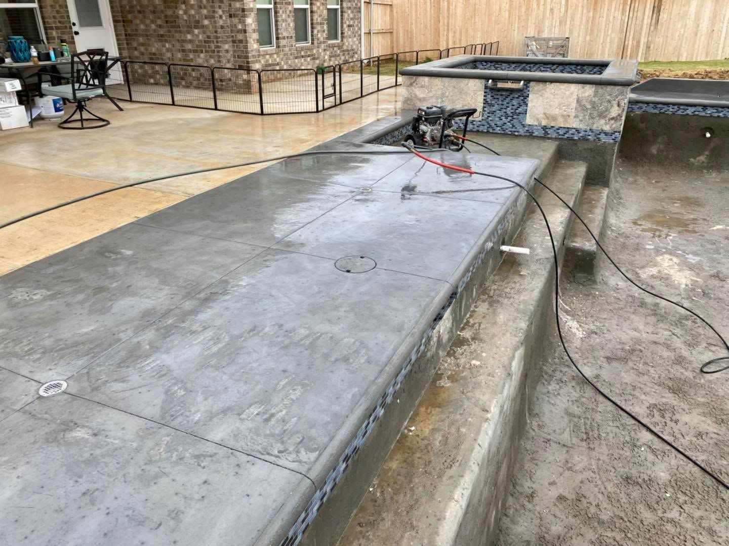 Wet concrete walkway beside a stone building and low wall, with hoses and a power tool on the surface.