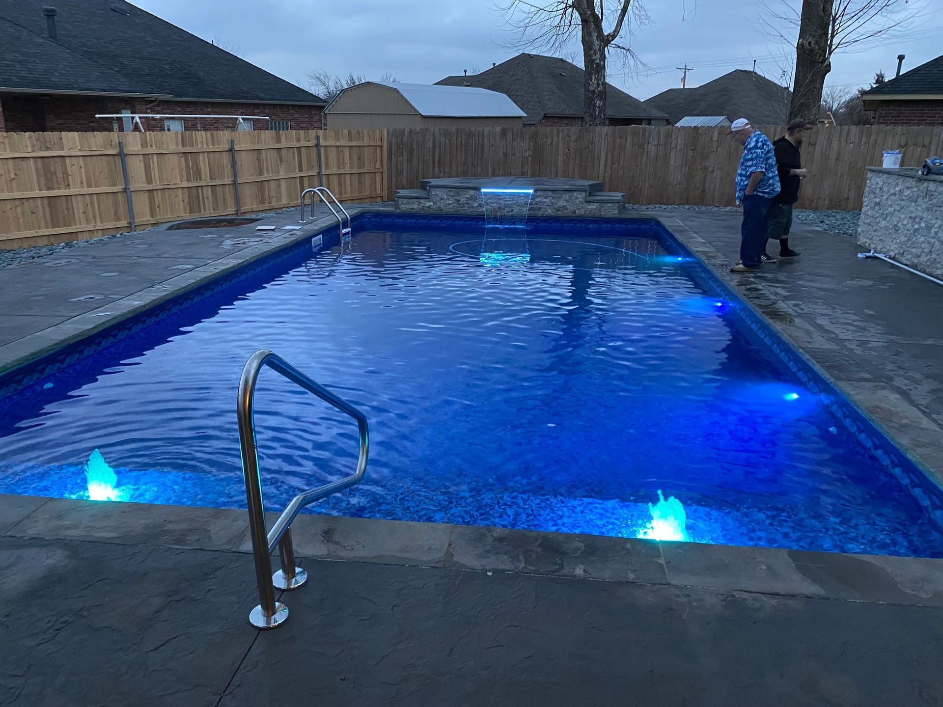 Rectangular backyard pool with blue underwater lights, a handrail, fence, and a person standing at the far end.