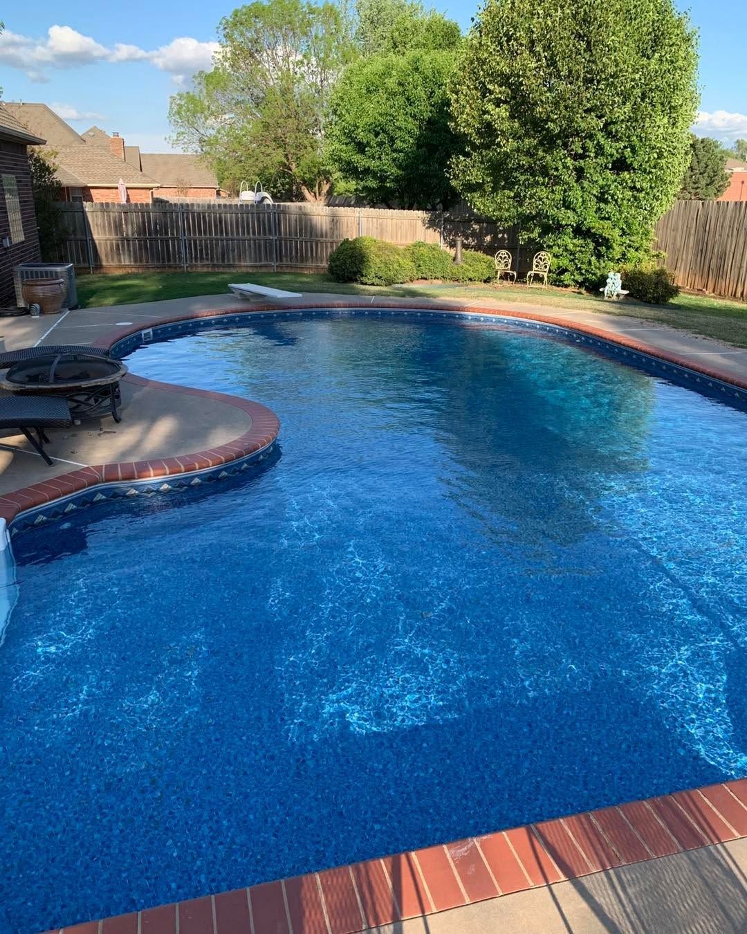 Backyard swimming pool with blue water, red brick border, lounge chair, and trees in the background