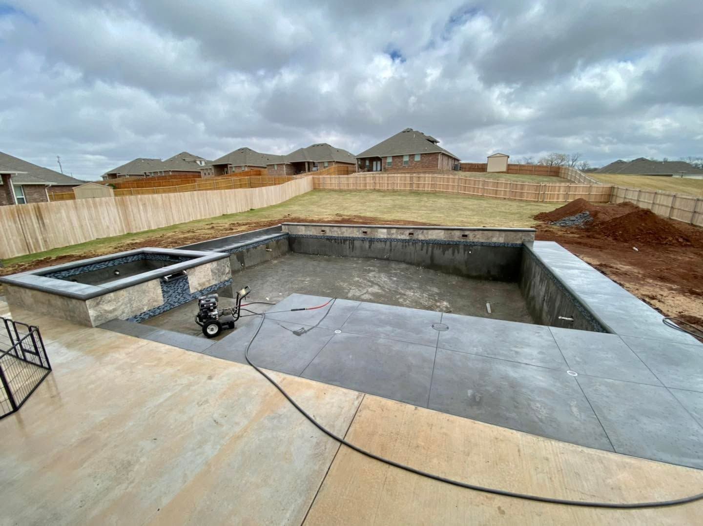 Backyard swimming pool under construction with concrete deck, fencing, and cloudy sky
