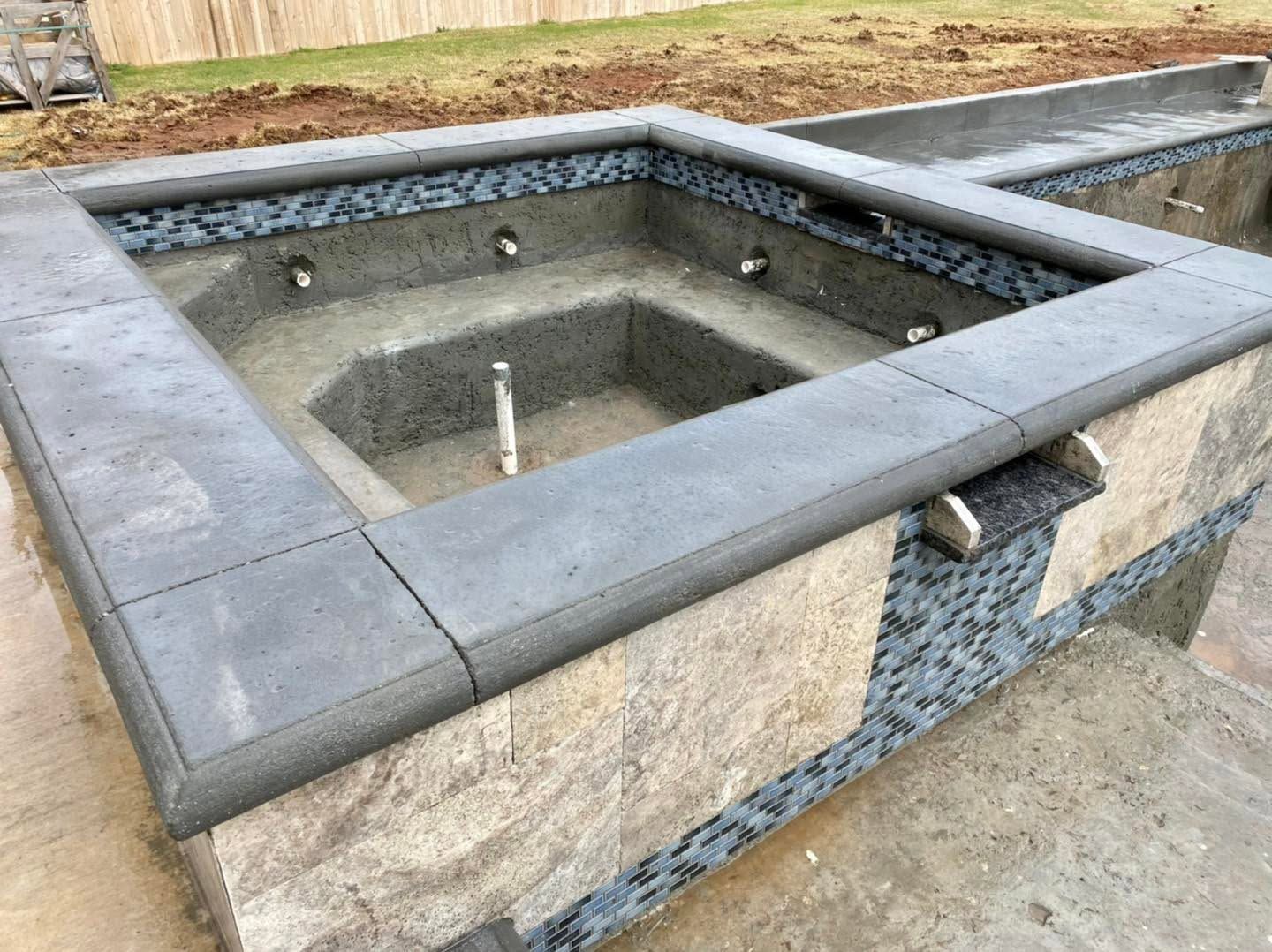 Outdoor stone hot tub or spa basin with gray tile coping and unfinished interior near a wooden fence