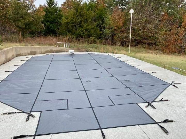 Blue pool cover stretched over an outdoor in-ground pool, secured with straps and surrounded by concrete decking.