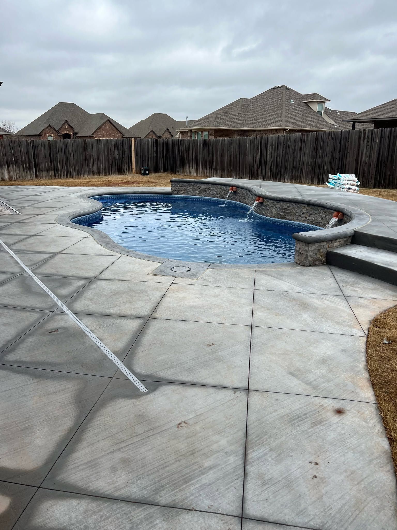 Backyard pool with cracked concrete deck, rock waterfall, and wooden privacy fence under cloudy sky