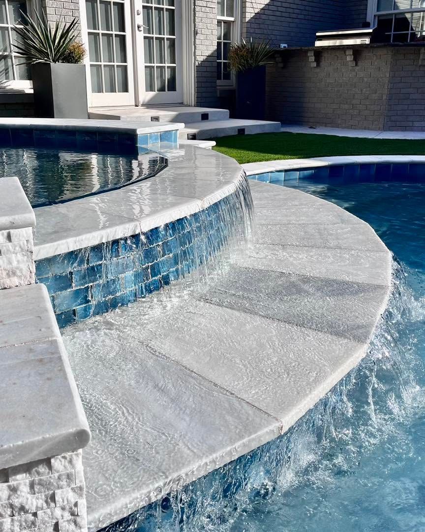 Curved stone steps leading into a blue swimming pool beside a tiled spa with water cascading nearby