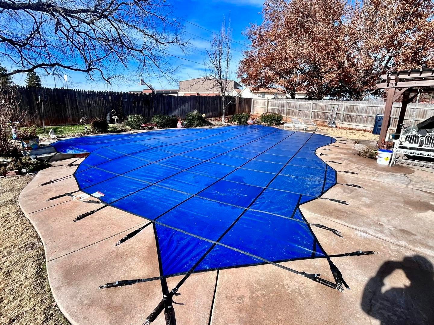 Blue tarp covering a backyard pool, surrounded by a concrete patio and leafless trees.
