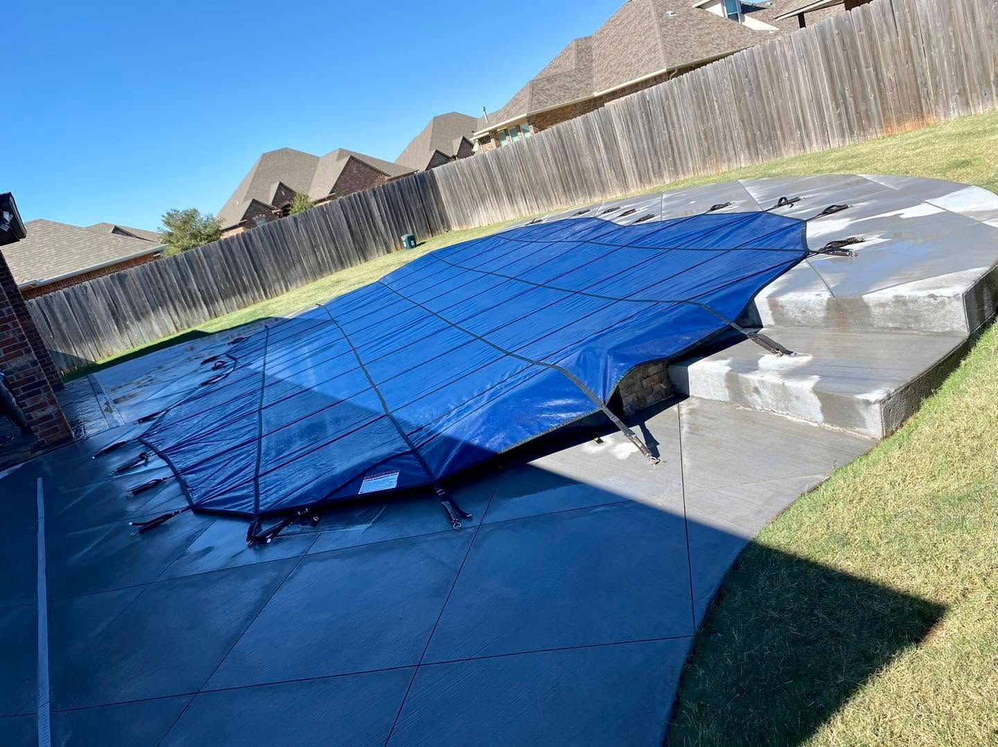 Blue pool cover stretched over an in-ground backyard pool beside gray steps and a wooden fence.