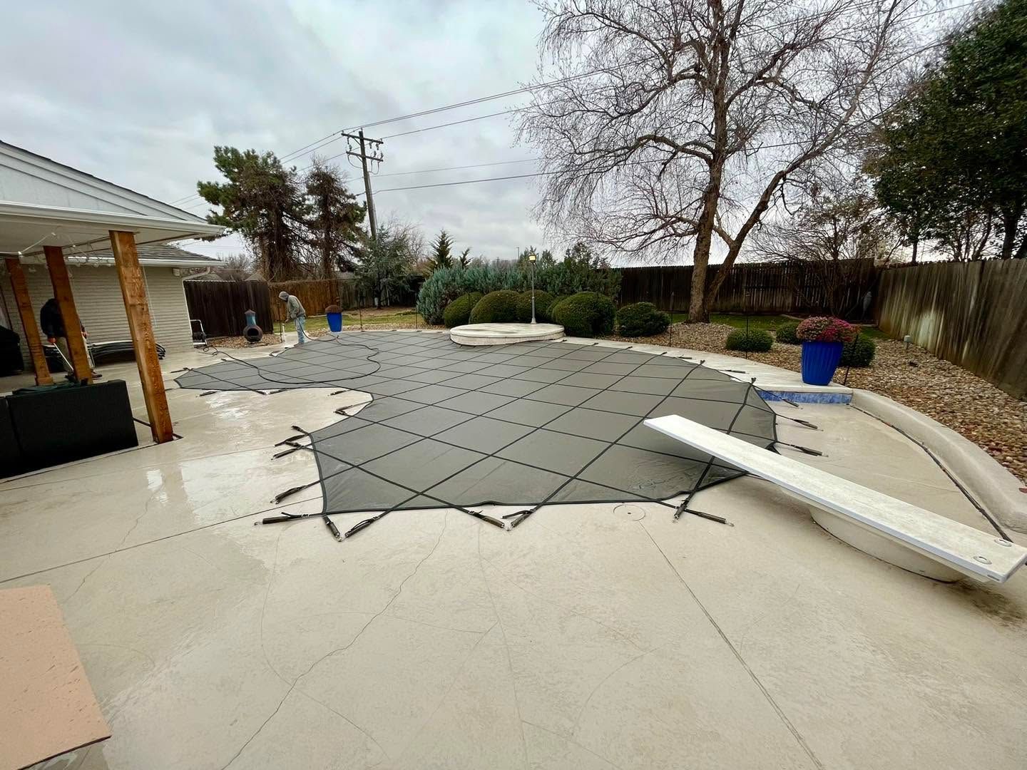 Backyard patio with torn pool cover, patio furniture, and trees under an overcast sky
