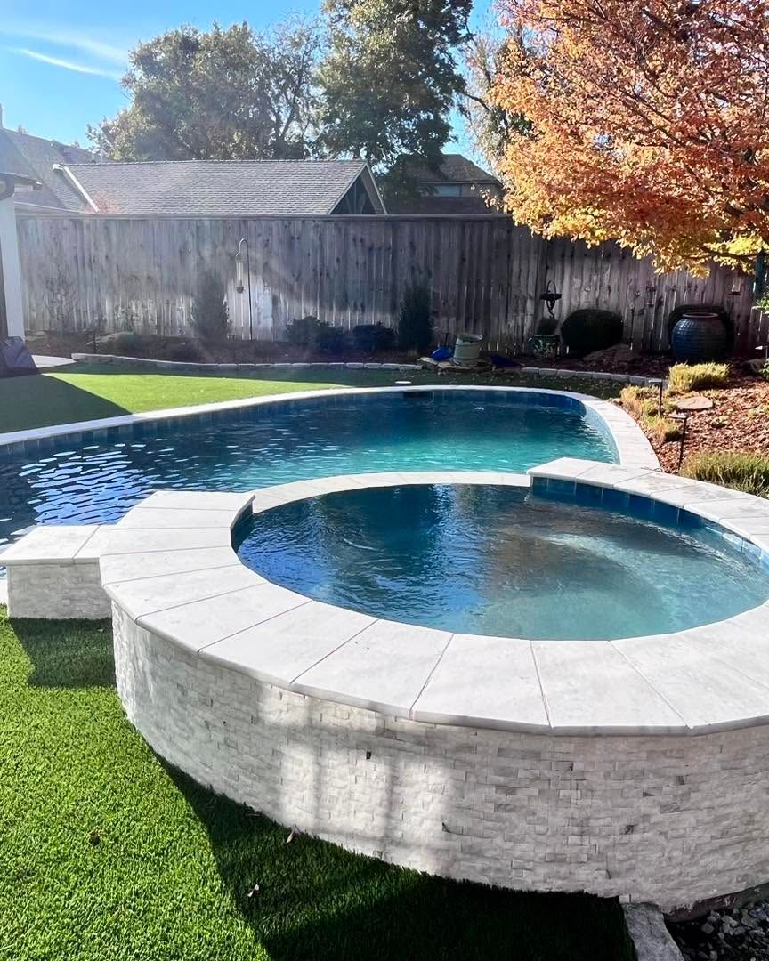 Backyard with a pool and round hot tub, bordered by green turf and autumn trees.
