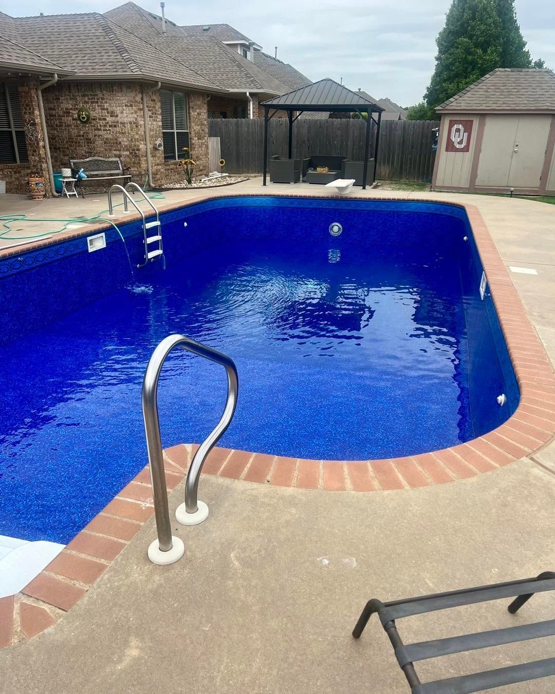 Backyard swimming pool with bright blue water, brick edging, and a metal handrail in the foreground