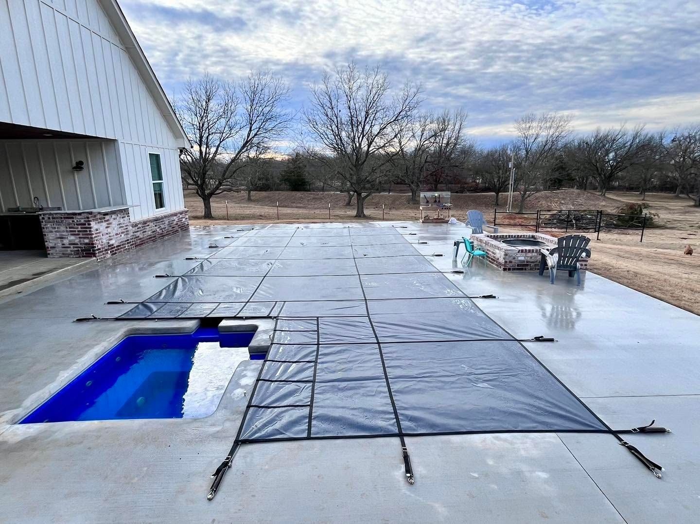 Backyard patio with a blue pool, covered in black protective sheeting, beside a house under a cloudy sky