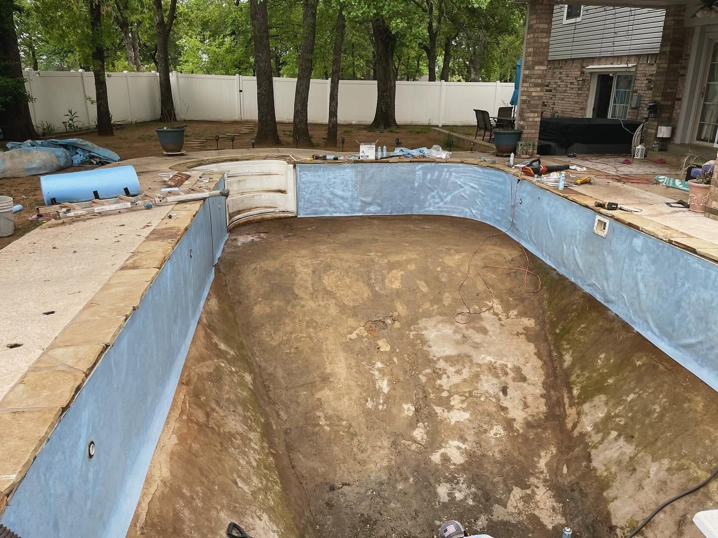 Empty backyard pool under construction with blue-painted walls and a dirt floor, surrounded by trees and patio tools