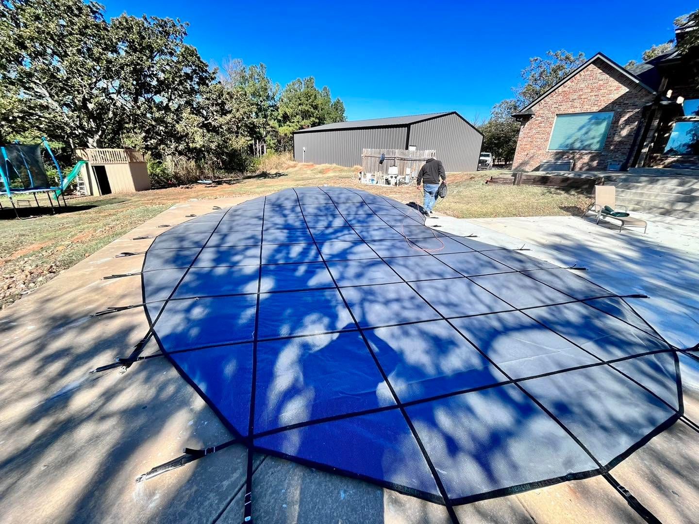 Large blue-and-white tarp or mat spread across a sunlit yard with trees, sheds, and a person in the background