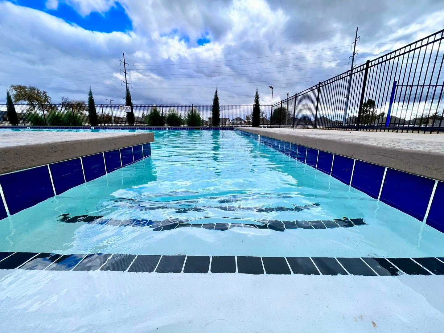 Blue-tiled outdoor pool with calm water, fence, and cloudy sky