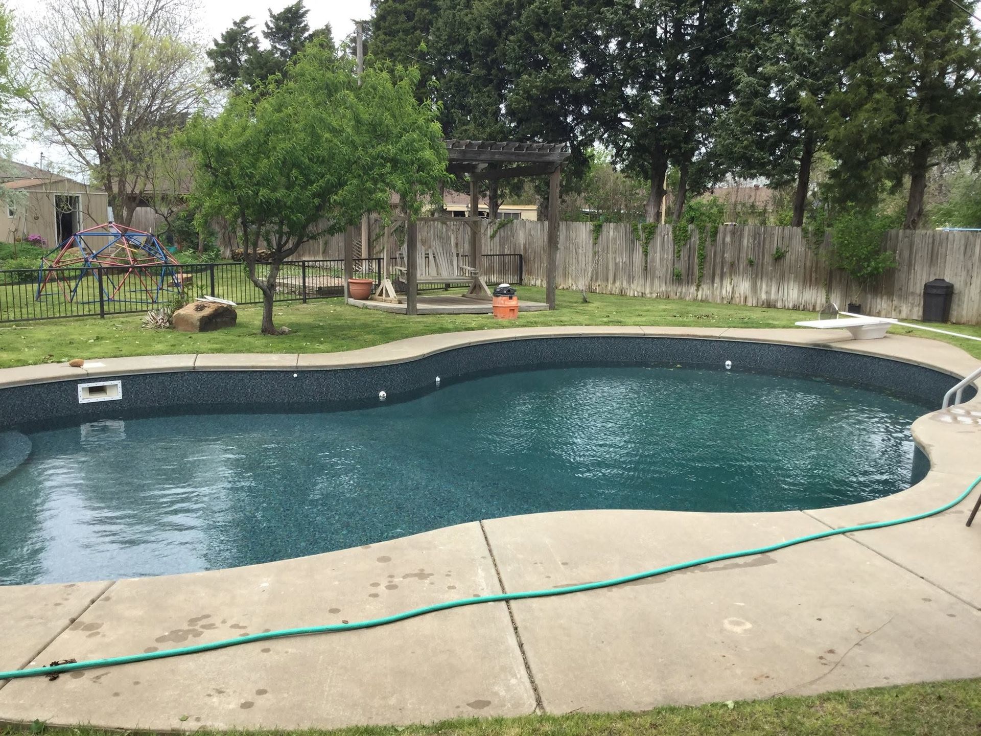 Backyard swimming pool with a green hose on the concrete deck and trees in the background