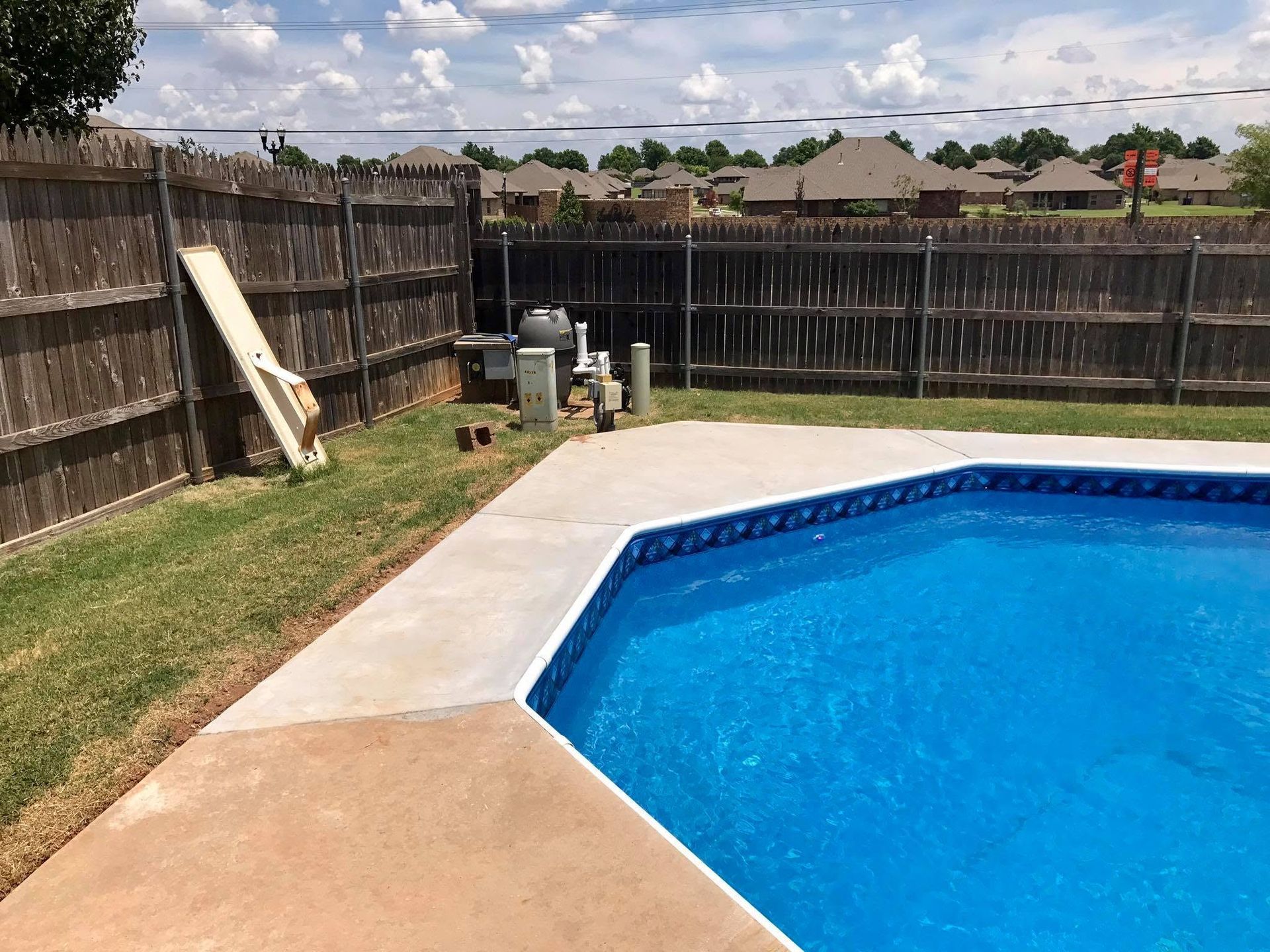 Backyard swimming pool with blue water, concrete deck, fence, and cloudy sky