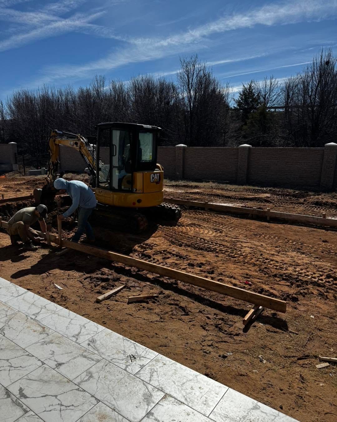 Worker using a power saw beside a yellow excavator on a dirt construction site under a blue sky