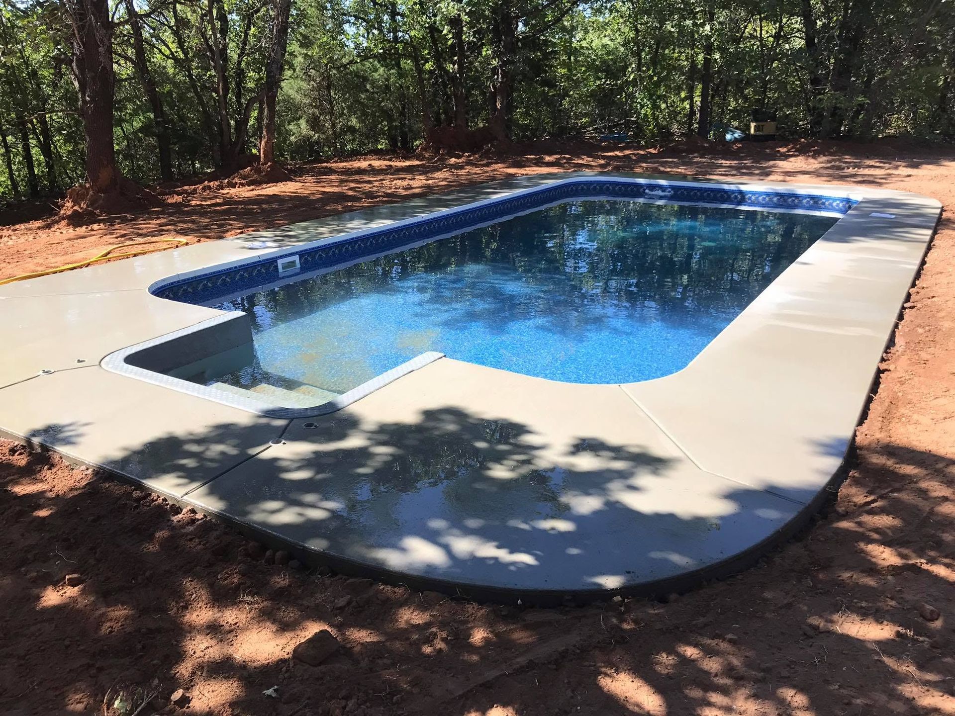 In-ground swimming pool with blue water surrounded by concrete in a wooded backyard